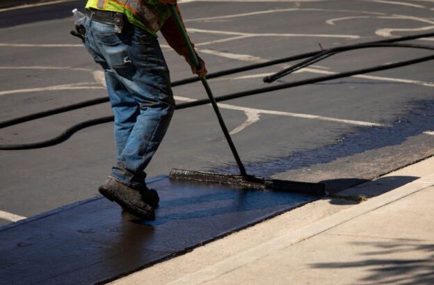 A person with a reflective yellow jacket resealing an asphalt driveway.