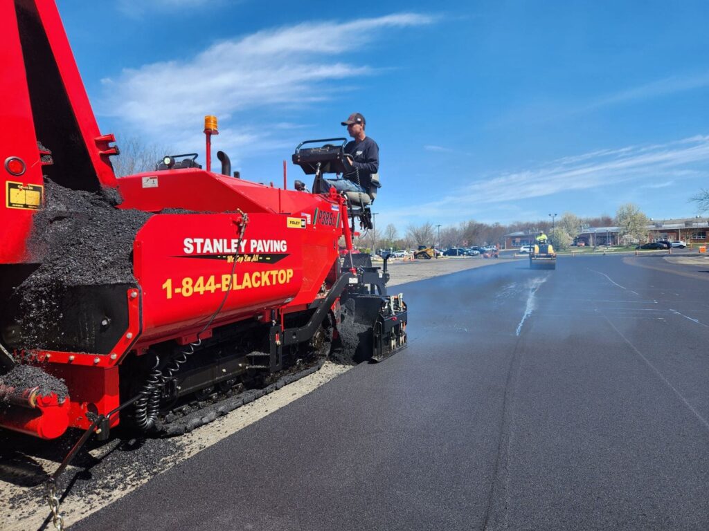 Stanley Paving crew operating a red asphalt paving machine while resurfacing a large parking lot under a clear blue sky. A roller compactor smooths the fresh asphalt in the background. The machine is branded with the company name and 1-844-BLACKTOP.