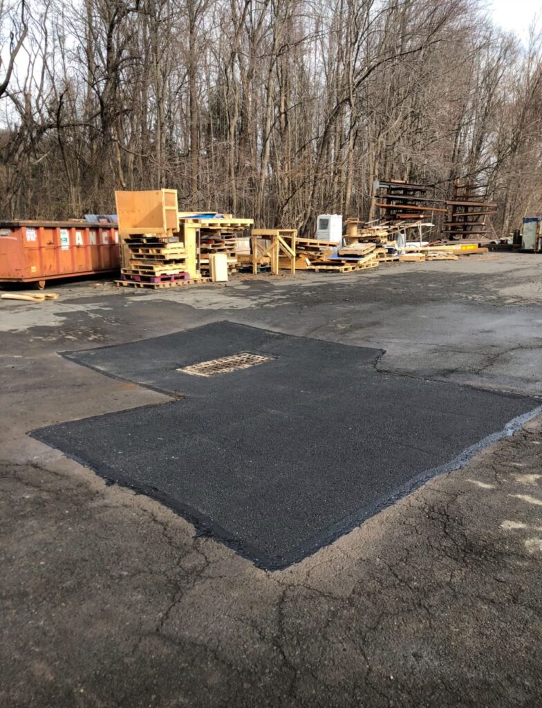 Freshly patched section of asphalt surrounding a drainage grate in an industrial yard, with old cracked pavement and stacked wooden pallets in the background.
