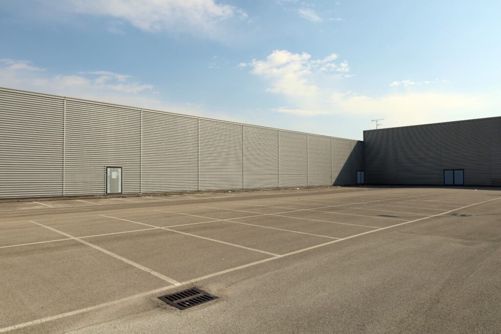 Empty commercial parking lot with freshly painted white line striping, bordered by a modern industrial building under a clear blue sky.