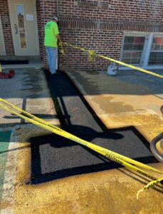 Stanley Paving crew member applies caution tape around a freshly patched asphalt walkway outside a brick commercial building, ensuring the area remains undisturbed while the surface cures.