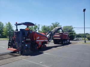 A Stanley Paving crew member operates a red asphalt milling machine in a large parking lot, loading removed pavement into a red dump truck marked with the Stanley Paving logo under clear blue skies, with trees and power lines in the background.