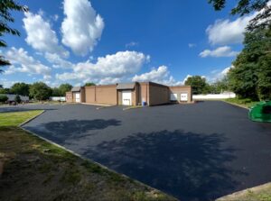 Newly paved commercial asphalt lot with a smooth blacktop finish outside a brick warehouse building in New Jersey, under a bright blue sky with scattered clouds.