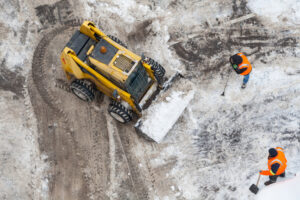 Aerial view of a yellow skid-steer loader and two workers in orange safety vests removing snow from a commercial parking lot.