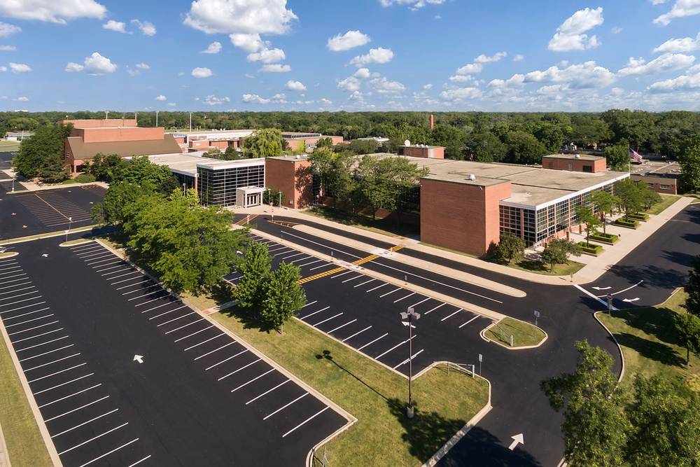 Aerial view of a large brick institutional building with a newly paved asphalt parking lot, clearly marked parking spaces, drive lanes, landscaped islands with trees, and surrounding greenery under a blue sky with scattered clouds.