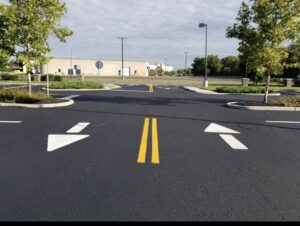 Freshly paved commercial parking lot with smooth asphalt, bright yellow center lines, and directional arrows leading toward an intersection, surrounded by curbs, landscaped islands, and light poles.