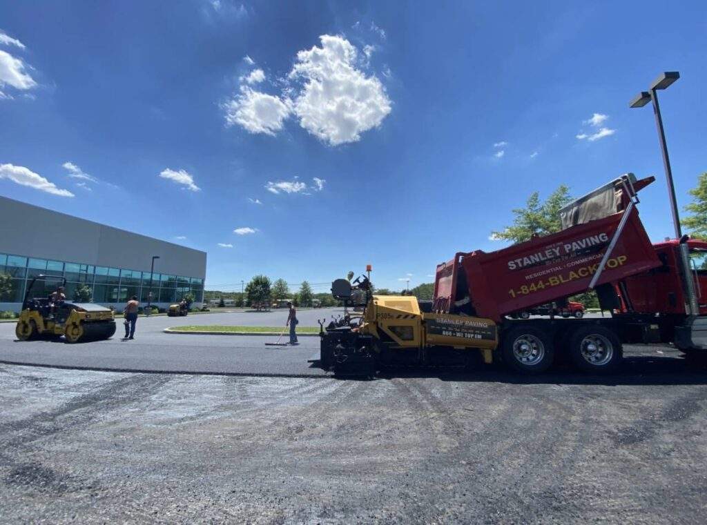Asphalt paving crew working on a commercial parking lot, with a paving machine, dump truck, and roller compactors under a clear blue sky.