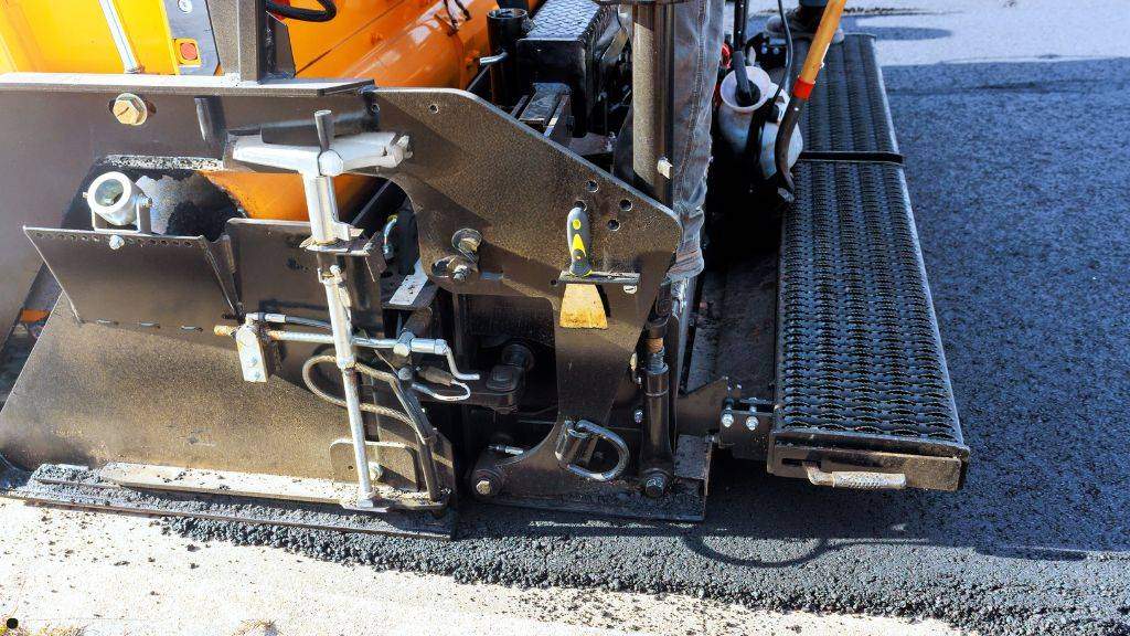 Close-up view of an asphalt paver screed laying a smooth new hot-mix blacktop surface over a milled parking lot before winter. 